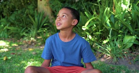 Preteen boy sitting cross-legged on grass looking up in sunlit garden wearing blue shirt