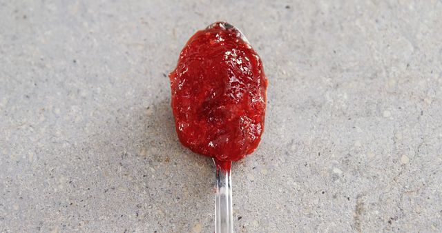 A spoonful of red jam rests on a concrete surface, with copy space. Its vibrant color and texture stand out against the gray background, suggesting a homemade or artisanal quality.