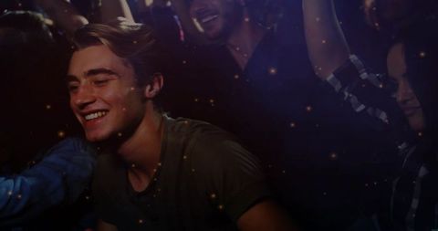 Smiling young man leaning into nightclub crowd with warm bokeh lights and haze, energetic celebratio