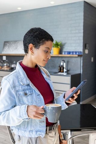 African american woman checking smartphone while holding latte in contemporary kitchen