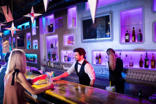 Bartender in trendy bar serving cocktails to customers at illuminated bar counter. Ideal for use in articles about nightlife, social scenes, bars, and hospitality industry. Suitable for marketing materials for bars, restaurants, and nightlife venues.