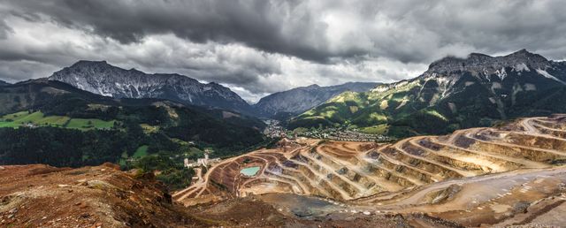 Panoramic view of an open-pit mine set against majestic mountain scenery under a dramatic cloudy sky. Green valleys contrast with the earthy tones of the mine, creating a striking juxtaposition of industry and nature. This stock photo is suitable for topics related to mining, environmental impact, natural landscapes, and industrial activities.