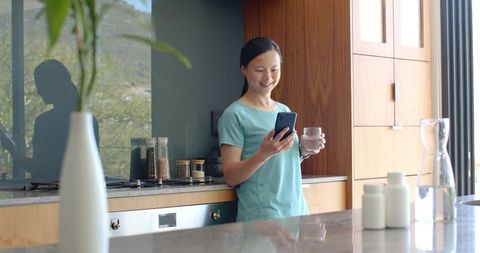 Asian woman leaning at kitchen counter, smiling while checking phone and holding glass