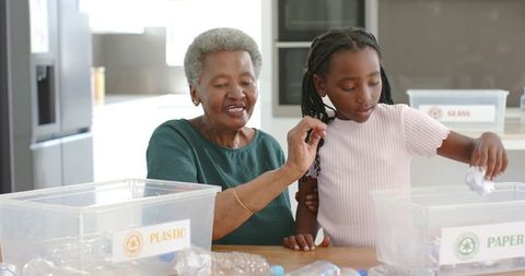 Grandmother teaching granddaughter recycling at kitchen table, sorting plastic and paper