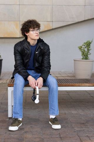 Young man sitting on bench wearing leather jacket holding headphones looking thoughtful