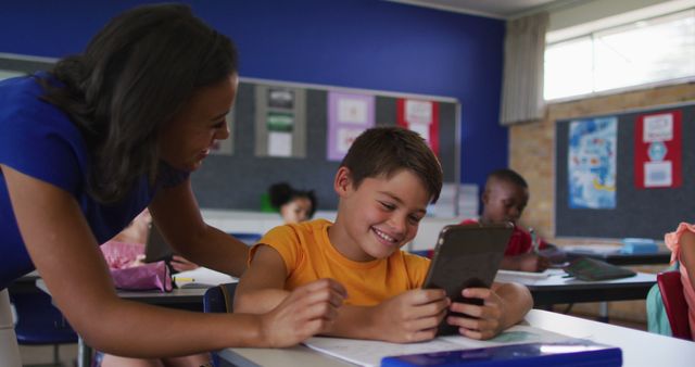 Teacher assisting a smiling boy using a tablet in a classroom with other students in the background. Useful for illustrating modern educational techniques, classroom engagement, digital learning, and teacher-student interactions.