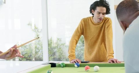 Multiracial friends leaning over pool table checking shot and smartphone during game