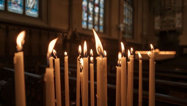 Warm glowing taper candles in historic chapel lighting wooden pews and stained glass glow