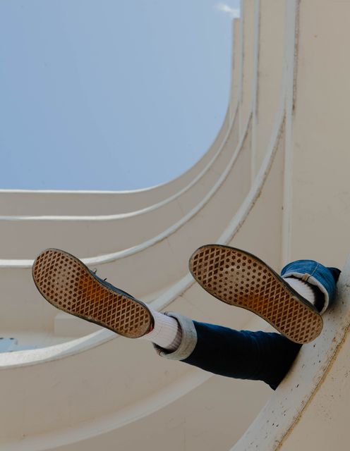 This image shows a person dangling their legs out of a modern architectural building. The focus is on the legs and the soles of their casual shoes, with a clear contrast between the smooth, abstract lines of the building and the person’s clothing. Use this as a stylish representation of modern urban life, a creative perspective on architecture, or a metaphor for relaxation and unconventional lifestyles.