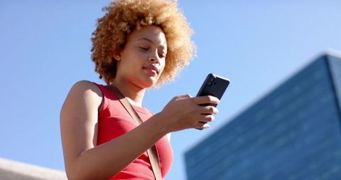 Young woman checking smartphone on urban steps wearing red top by glass building in daylight