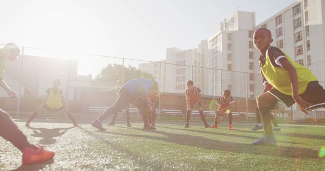 Boys in soccer uniforms engaging in warm-up exercises on a sunny urban soccer field. Ideal for use in educational materials, community sports promotion, physical fitness campaigns, and advertisements for youth sports programs.