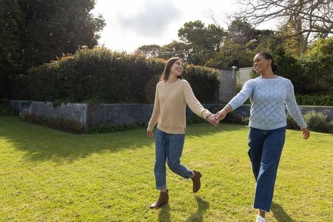 Mixed-race female partners walking hand in hand on green lawn, smiling in casual sweaters