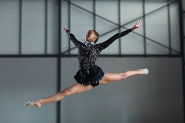 Biracial female gymnast wearing a gymnastics costume practicing at the gym, jumping and doing splits in the air with her arms raised. Gymnast training hard for competition.