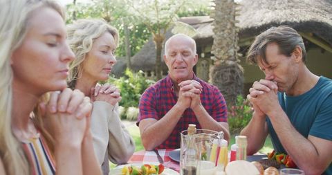 Caucasian family of four sitting at an outdoor table, praying together before meal. Surrounded by lush greenery, evoking atmosphere of unity and gratitude. Ideal for themes of family values, unity, thanksgiving celebrations and serene outdoor living.