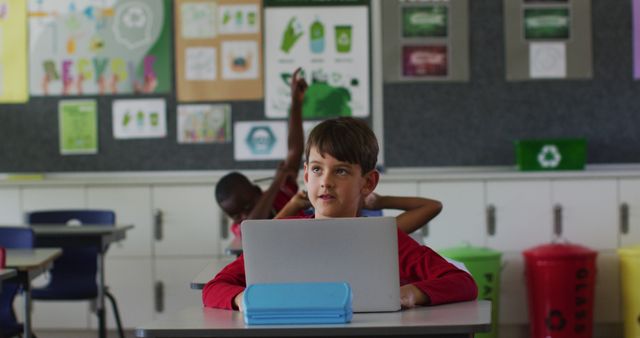 Young boy in a classroom focused on using a laptop with environmental posters on walls and recycling bins in the background. Ideal for concepts related to education, environmental awareness, sustainability studies, and classroom activities.