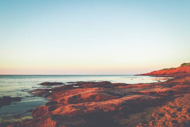 The setting sun casts a warm glow over the rocky beach, creating a tranquil scene perfect for promoting relaxation and natural beauty. This image is ideal for travel brochures, websites featuring coastal destinations, desktop backgrounds, and nature-focused blogs.