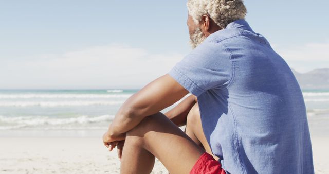 Senior african american man sitting and looking away on sunny beach. healthy and active time beach holiday.