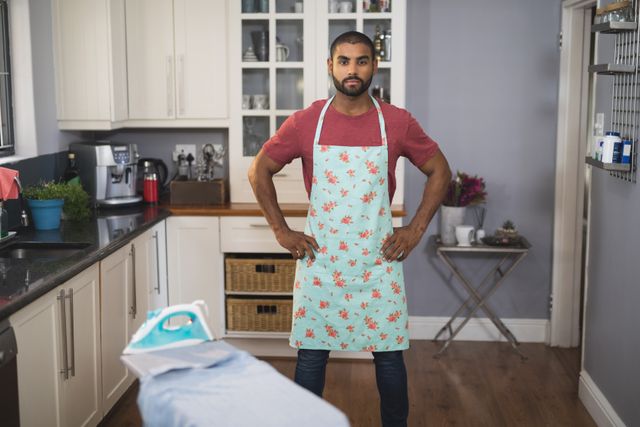 Young man standing confidently in a modern kitchen, wearing a floral apron. Ideal for use in articles or advertisements related to home cooking, lifestyle, domestic chores, or modern kitchen designs.