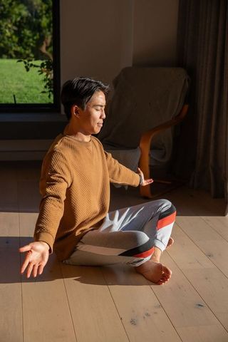 Sunlit asian man meditating cross-legged at home on wooden floor, cozy minimalist livingroom