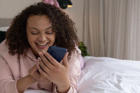African american woman relaxing on bed smiling while using smartphone, cozy pastel bedroom