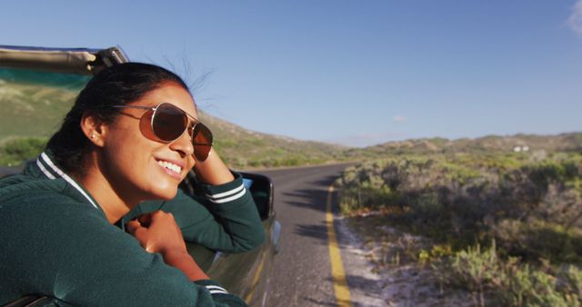 Woman leaning out car window happily enjoying road trip on clear sunny day with scenic nature view. Perfect for concepts related to travel, freedom, adventure, outdoor activities, and happiness.