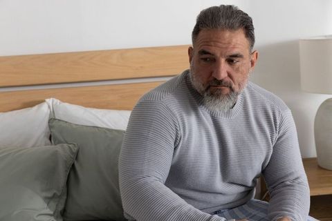Mature man sitting on bed edge looking contemplative in cozy neutral bedroom