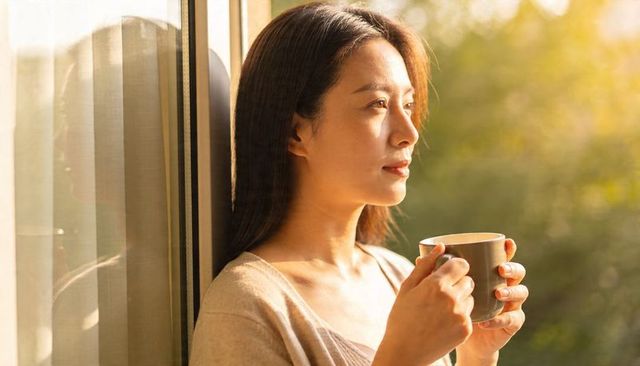 Asian woman enjoying morning coffee by sunlit window, contemplative golden hour moment