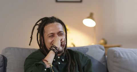 Joyful african american man laughing on sofa covering mouth bracelets, cozy living room
