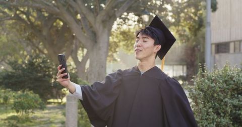 Asian male graduate taking selfie on sunlit university quad in cap and gown