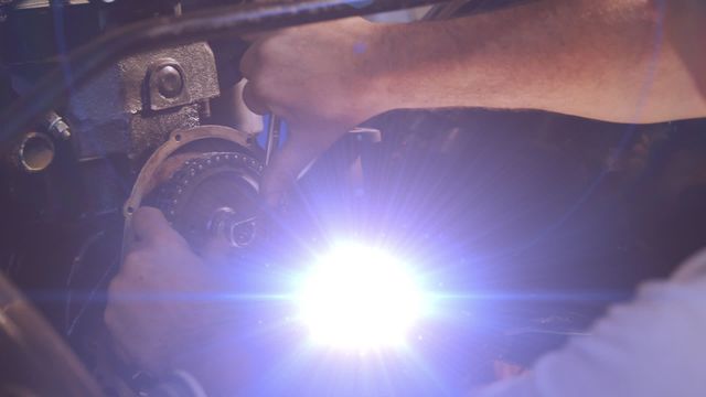 Close-up view of mechanic working on car engine with a bright light illuminating the area. Useful for automotive repair content, vocational training materials, and automotive service advertisements.