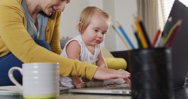 Caucasian mother holding her baby using laptop and talking on smartphone while working from home. motherhood, love and childcare concept