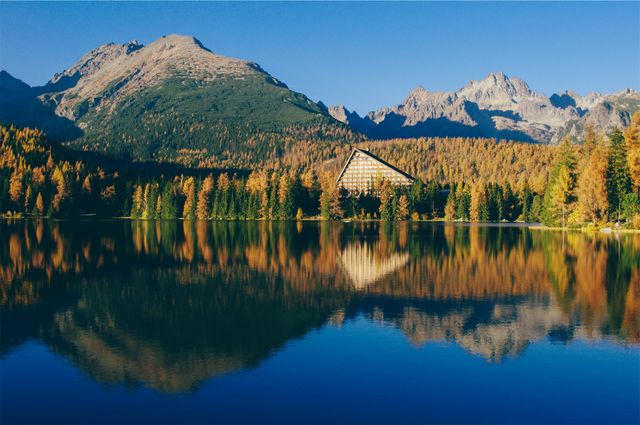 Beautiful mountain lake surrounded by autumn-colored trees with a modern pyramid-shaped hotel reflecting on the water. The serene landscape with clear blue skies makes an ideal backdrop for travel promotions, tourism ads, nature magazine covers, or scenic calendars.