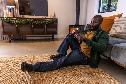 African american man lounging on rug, scrolling smartphone by cozy fireplace and garland