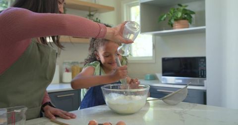 Multicultural mother and daughter whisking batter on kitchen island while pouring milk