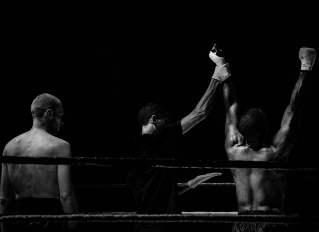 Black and white photo capturing a moment of victory in a boxing match. The victorious boxer is seen with arms raised triumphantly, while the referee lifts his hand to declare him winner. Another competitor is present with a disappointed demeanor. Strong dramatic lighting and contrast emphasize the intensity and emotion of the moment. Ideal for use in sports promotions, motivational posters, and advertisements related to fitness and competition.