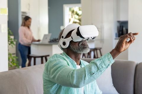 Senior man using virtual reality headset at home while partner working on laptop nearby