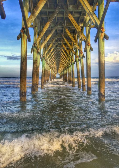 Underneath wooden pier with ocean waves gently crashing against wooden poles during sunset. Perfect for themes of tranquility, architecture, beachfront scenes, seaside vacations, and marine life. Useful for travel magazines, coastal tourism advertisements, or calming oceanic backgrounds for websites and presentations.