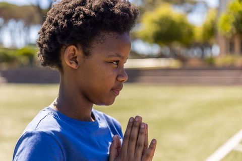 African american schoolboy praying on sunlit field wearing blue tee, serene moment