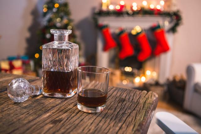 This image shows a close-up of a bottle of whiskey and a glass on a rustic wooden table. In the background, a fireplace is adorned with festive Christmas decorations, including stockings and lights. This image can be used for holiday-themed advertisements, social media posts, or articles about festive drinks and cozy celebrations.