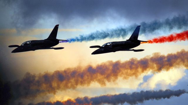 Jets performing aerobatic stunts with colorful smoke trails during sunset capture the thrilling atmosphere of an air show. Ideal for use in aviation-themed publications, marketing materials for air festivals, and as part of designs highlighting precision flying and aerial acrobatics.
