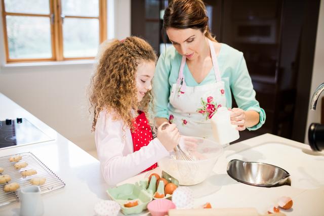 Mother and daughter are seen baking together in a bright, modern kitchen. The mother is assisting her daughter who is whisking flour in a mixing bowl. Both of them are wearing aprons, indicating they are involved in a cooking activity. This image portrays family bonding, joy, and togetherness. Ideal for advertisements and articles on parenting, family activities, cooking recipes, or kitchen products.