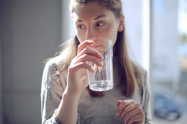 Woman drinking water in a room filled with natural sunlight, captured in a moment of thoughtfulness. Represents healthy lifestyle, hydration, and morning routines. Ideal for health and wellness campaigns, advertisements for water or healthy living products, and articles promoting self-care and hydration.