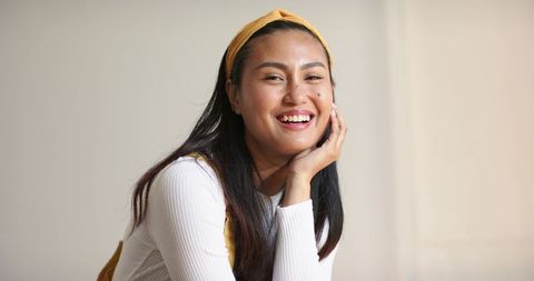 Young Asian woman smiling, resting hand on cheek, wearing yellow headband and white top