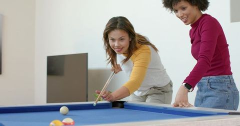 Diverse friends playing pool in modern home living room, leaning over blue felt table
