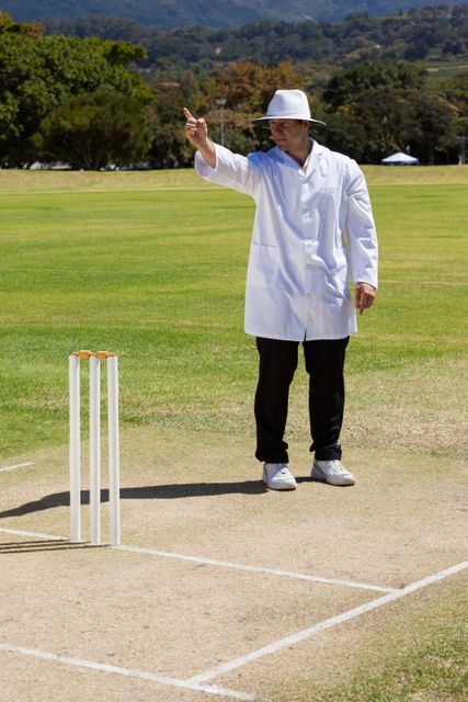 Cricket umpire in white coat and hat signalling out during a match on a sunny day. Ideal for use in sports-related content, cricket training materials, articles about cricket rules, or promotional materials for cricket events.