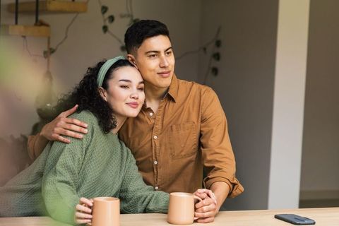 Young couple enjoying cozy morning coffee at home, cuddling and looking out window