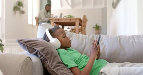 African american father working from home while son relaxing with headphones on sofa