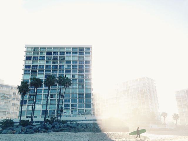 Surfer with board walking on sandy beach at sunrise with modern towers in background and palm trees on side. Soft mist and light reflection create tranquil, serene atmosphere. Ideal for themes of urban coastal life, leisure activities, morning routines, and nature meeting city.