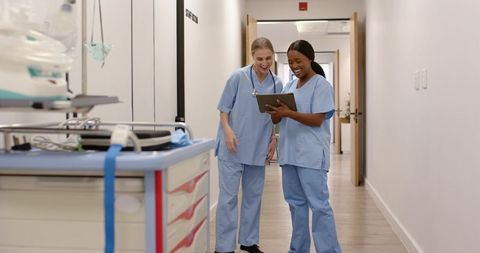 Smiling nurses reviewing tablet while checking crash cart in hospital corridor