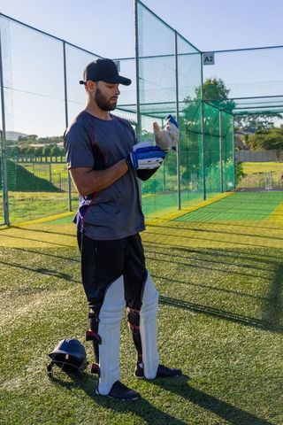 Cricket player preparing batting gear in sunlit practice nets on synthetic turf